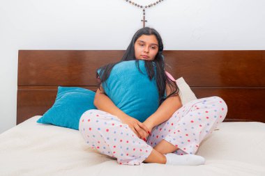 Yatakta oturan genç Latin bir kadın yastığı göğsüne sıkıca bastırıyor. Gözleri düşüncelere dalmış ve ifadesi derin üzüntü ve duygusal acıyı yansıtıyor..