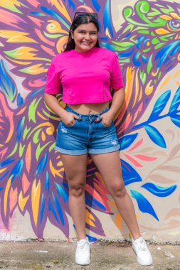 Pembe tişörtlü ve kot şortlu şık Latin bir kadın. Renkli bir grafiti duvarının önünde kafasına güneş gözlüğü takıp moda modasına uygun poz veriyor..