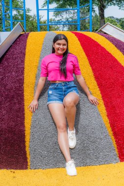 Pembe bluzlu ve kot şortlu gülümseyen Latin kadın renkli bir kaydırak üzerinde poz veriyor. Neşeli bir açık hava ortamında tüm vücut portresi.