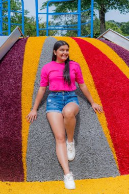 Pembe bluzlu ve kot şortlu genç Latin kadın renkli bir kaydırağın üzerinde oturuyor, sağa bakıyor ve arka planda bir park ayarını görebiliyor..