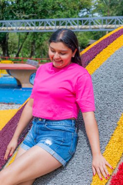 Pembe tişörtlü ve kot şortlu Latin bir kadının yarı vücut portresi. Bir bacağı açık havada canlı bir oyun alanında rahat ve düşünceli bir poz verirken vurgulanmış.