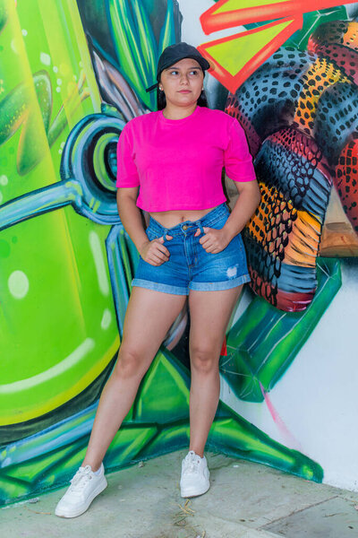 Полнотелый портрет стильной латиноамериканской женщины в черной шапке, розовом топе, джинсовых шортах и белых кроссовках, позирующей модно и смотрящей в сторону перед граффити-артом.