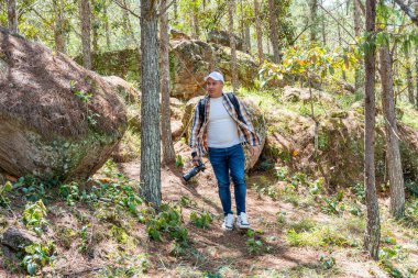 Latin Amerikalı fotoğrafçı hızla ormanda yürüyor, ağaçlara doğru bakıyor. Açık hava aksiyon sahnesi macera, keşif ve doğa ve fotoğrafçılık ile dinamik bir bağlantı yansıtıyor..