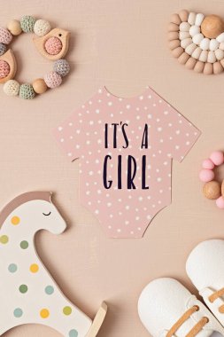 Baby shower, gender reveal party. It's a girl message over paper cut onesie. Flatlay, top view on a beige pastel background. Newborn gifts. Invitation, celebration, greeting card idea