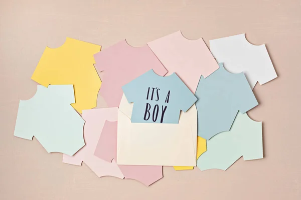 Baby shower, gender reveal party. It\'s a boy message over paper cut onesie. Flatlay, top view on a beige pastel background. Newborn gifts. Invitation, celebration, greeting card idea