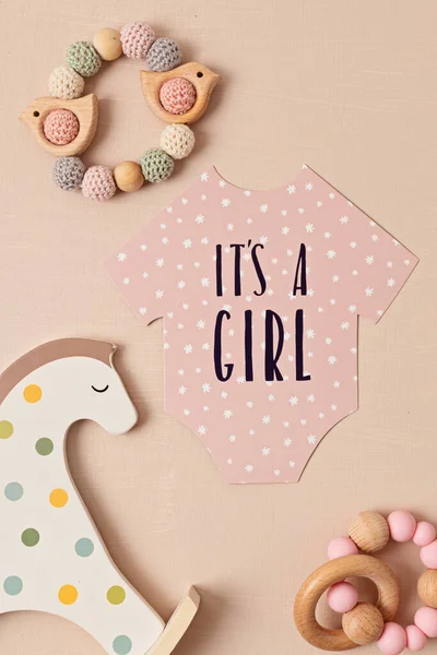 Baby shower, gender reveal party. It\'s a girl message over paper cut onesie. Flatlay, top view on a beige pastel background. Newborn gifts. Invitation, celebration, greeting card idea