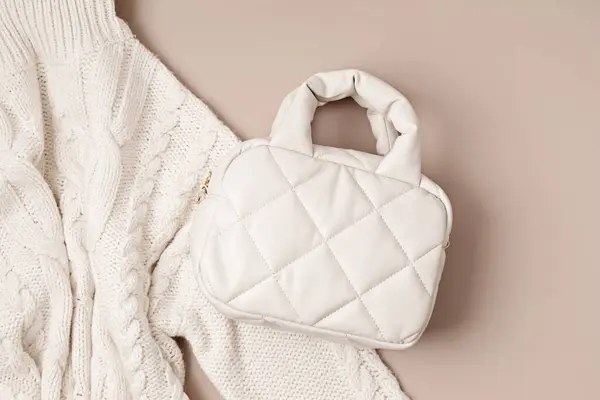 Beige quilted puffed bag and woolen sweater on pastel background. Stylish woman outerwear. Winter fashion accessories