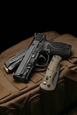 A pistol, cartridges for it and a folding knife on a brown backpack. Self-defense and survival kit. Compact edged weapons and firearms. Dark background.
