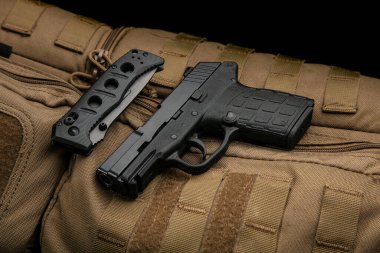 A small modern pistol and penknife on a brown military backpack. Self defense kit. Dark background.