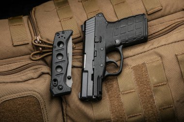 A small modern pistol and penknife on a brown military backpack. Self defense kit. Dark background.