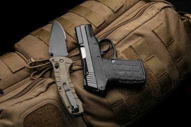 A small modern pistol and penknife on a brown military backpack. Self defense kit. Dark background.