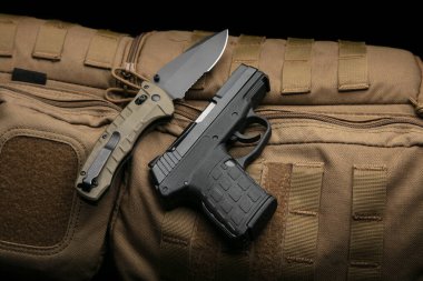 A small modern pistol and penknife on a brown military backpack. Self defense kit. Dark background.
