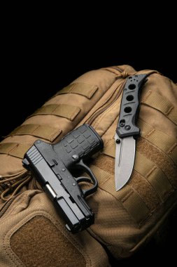 A small modern pistol and penknife on a brown military backpack. Self defense kit. Dark background.