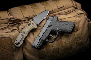 A small modern pistol and penknife on a brown military backpack. Self defense kit. Dark background.