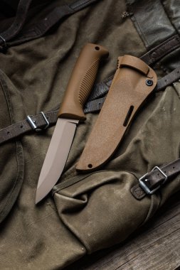 Lastik saplı ve plastik kılıflı modern bıçak. Soğuk çelik antika bir sırt çantasının üzerinde yatıyor. Ahşap arkaplan.