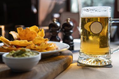 Soğuk bir kupa bira, bir tabak nachos ve tahtanın üzerinde bir kase guacamole 'nin yanında oturur, rahat ve davetkar bir yemek sahnesi yaratır, yiyecek ve içecek stoku fotoğrafları için mükemmeldir..