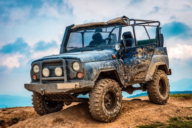 Çamurla kaplı engebeli bir off-road aracı, bulutlu bir gökyüzü zeminine karşı toprak bir tepeye park edilmiş, zorlu arazilerde dayanıklılığını ve kapasitesini sergiliyor..