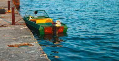 Küçük yeşil ve sarı bir motorlu tekne beton bir rıhtıma demirlemiş, etrafında sakin mavi bir su ve arkasında 25 etiketli bir dış motor var..