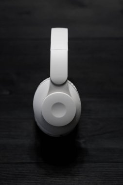 Bir çift beyaz kablosuz kulaklık, yastıklı kulaklıklar ve sol kulaklık üzerindeki kontrol düğmeleri, koyu ahşap bir yüzeye yerleştirilmiş..