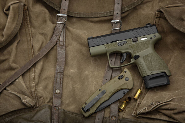 A tactical setup featuring a green and black handgun, a folding knife, and bullet casings on a brown military-style backpack, showcasing survival gear and outdoor preparedness.