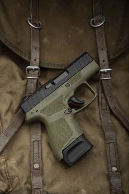 Deri kayışlı, engebeli kahverengi bir çantanın üzerine yerleştirilmiş yeşil ve siyah bir tabanca taktiksel ve dayanıklı bir estetik sergiliyor. Açık hava, askeri veya nefsi müdafaa teması için mükemmel..