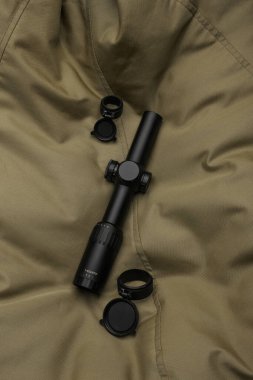 Profesyonel atıcılık ve açık hava kullanımı için hassasiyet, dayanıklılık ve taktik tasarımı vurgulayan desenli haki kumaş üzerinde mercek kapaklı siyah bir tüfek dürbünü..