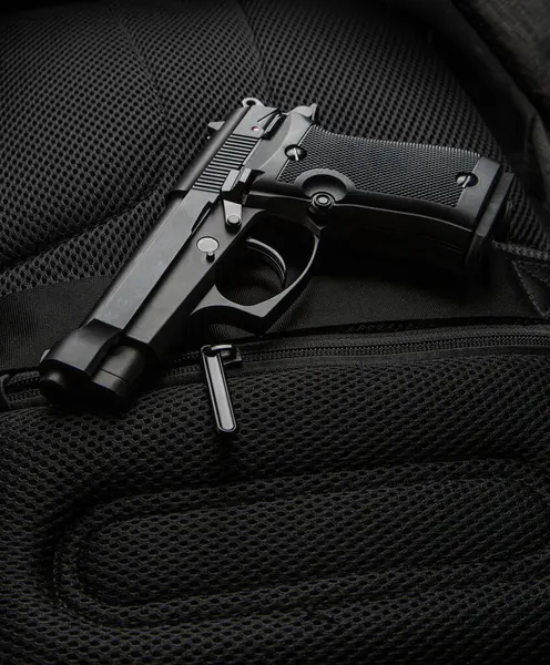 Siyah taktik tabanca, koyu renk bir sırt çantasının yastıklı ağ yüzeyine yerleştirilmiş. Yakın çekim, güçlü bir kontrast ve detay içeriyor.