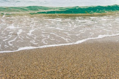 Küçük bir turkuaz dalga tepeye çıkar ve beyaz köpüğe dönüşür. Kaba, kahverengi kum üzerinde yıkanır.