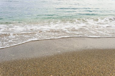 Küçük bir turkuaz dalga tepeye çıkar ve beyaz köpüğe dönüşür. Kaba, kahverengi kum üzerinde yıkanır.