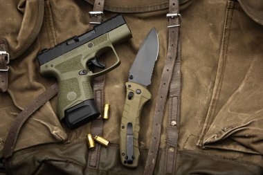 Tactical lay flat of an olive drab pistol, folding knife, and 9mm rounds on canvas.