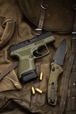 Tactical lay flat of an olive drab pistol, folding knife, and 9mm rounds on canvas.