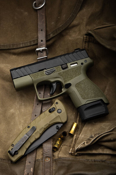 Tactical lay flat of an olive drab pistol, folding knife, and 9mm rounds on canvas.