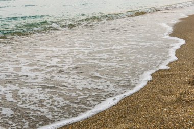 Küçük bir turkuaz dalga tepeye çıkar ve beyaz köpüğe dönüşür. Kaba, kahverengi kum üzerinde yıkanır.