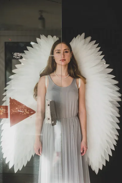 天使のイメージの少女 黒い髪の長い天使の女の子が 灰色のドレスと白い翼を持つガラスのドアのそばに立っている 肖像画 — ストック写真