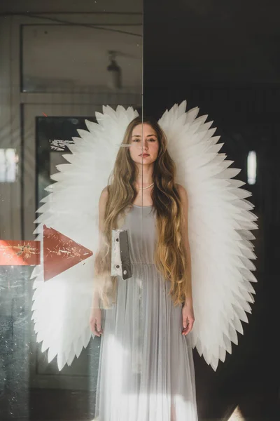 白い翼と灰色のドレスで長い暗い髪の天使の女の子は ガラスのドアのそばに立っている — ストック写真