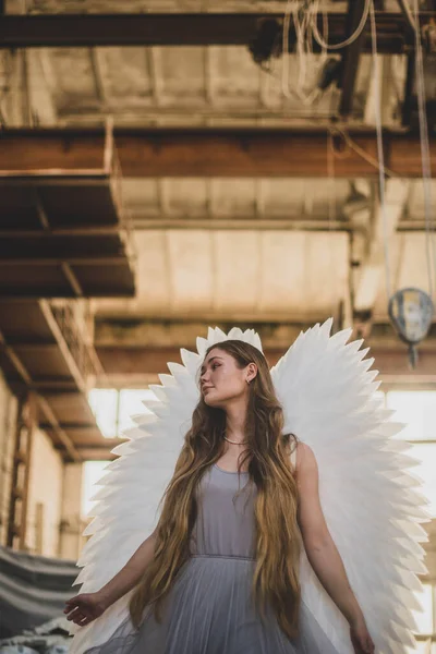 天使の少女が工房に立っている 彼女の後ろには大きな白い翼がある 女の子はワークショップの背景を背景に腕を伸ばして横を見る — ストック写真