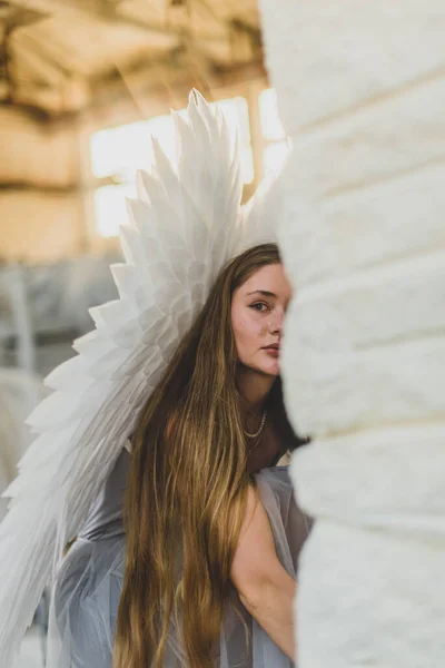 若い美しい天使の女性の肖像画 少女はワークショップに座って壁の後ろから外を見る 背中には大きな白い翼がある 優しい天使 — ストック写真
