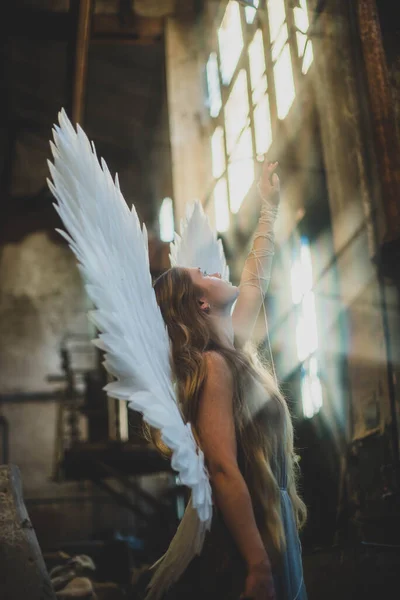 天使の少女は乱雑なワークショップに立っている 背中に翼を持つ女の子が太陽のために手を伸ばす 天使が見上げた 手に糸を — ストック写真