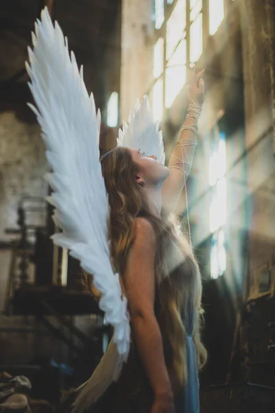 天使の少女は乱雑なワークショップに立っている 背中に翼を持つ女の子は太陽のために手を差し伸べ — ストック写真