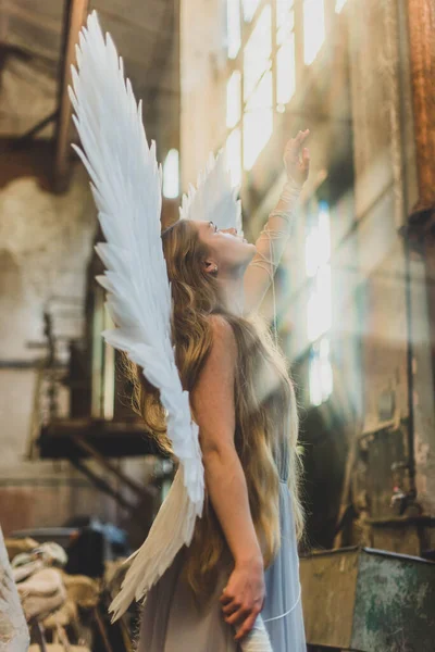天使の少女は乱雑なワークショップに立っている 背中に翼を持つ女の子は太陽のために手を差し伸べ — ストック写真