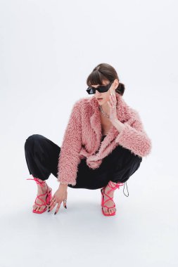 Fotoğraf stüdyosundaki muhteşem modelin tam boy fotoğrafı, şık pembe kürk manto, eşofman altı, güneş gözlüğü, topuklu ayakkabı. Moda, tarz