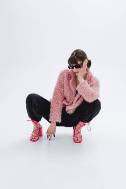 Fotoğraf stüdyosundaki muhteşem modelin tam boy fotoğrafı, şık pembe kürk manto, eşofman altı, güneş gözlüğü, topuklu ayakkabı. Moda, tarz