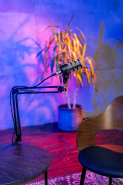 A vibrant and modern workspace features a microphone on an adjustable arm, an empty chair, and colorful lighting against a wall. This setup is ready for an in-depth podcast discussion. clipart