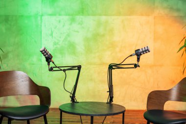 İki ayarlanabilir mikrofon canlı bir stüdyo ortamında bir yuvarlak masaya yerleştiriliyor ve podcast röportajı için davet edici bir alan yaratıyor. Arkaplanda parlak renkler var.