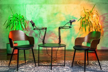 Profesyonel mikrofonlarla donatılmış bir masada birbirine bakan iki ahşap sandalye, hepsi canlı yeşil ve desenli arka plana karşı kurulmuş. Sıcak atmosfer canlı bir podcast oturumu öneriyor.