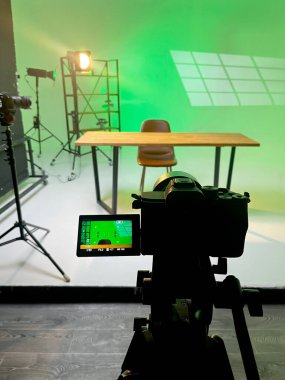 Podcast 'te mikrofonlar ve stüdyo ışıklarıyla çevrili ahşap bir masa ve sandalye yer alıyor. Yeşil bir arkaplan kayıt ortamını güçlendirir.