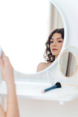 Uzun, dalgalı saçlı genç bir kadın dairesel bir aynada makyaj uygulamasına odaklanıyor. Oda doğal ışıkla yıkanır. Hazırlanırken yüzünün rengini gösterir..