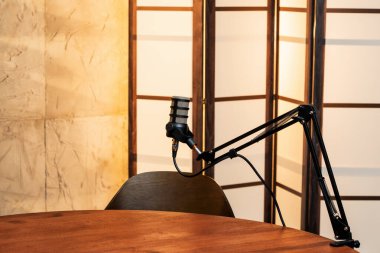 İyi donanımlı bir podcast stüdyosunda, ayarlanabilir bir kolda sıcak ve davetkar bir arkaplana karşı ayarlanmış bir mikrofon bulunur. Tahta bir masa ve boş bir sandalye yaklaşan tartışmalara ipucu veriyor..