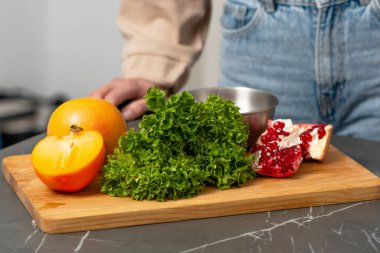 A wooden cutting board displays vibrant fresh ingredients including pomegranate, crisp green lettuce, and ripe persimmon. A blurred figure stands nearby in a stylish kitchen, adding a homey touch. clipart