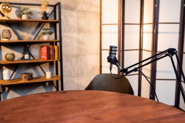 İyi düzenlenmiş bir podcast alanı, dekoratif eşyalar ve sıcak ışıklandırmalarla çevrili, kayda hazır ahşap bir masa ve şık bir mikrofon sunar..